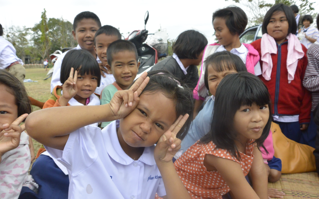A group of Thai schoolchildren smiling and playfully posing for the camera outdoors.